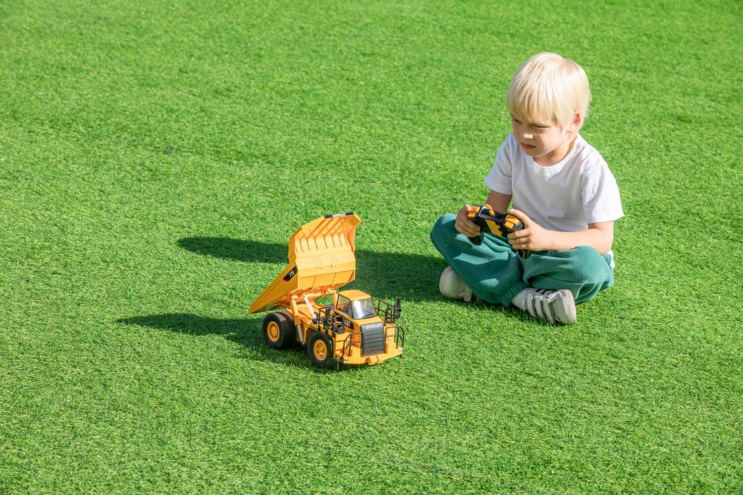 Blonde child seated on grass operating a remote control with a toy dump truck in front, demonstrating product photography.