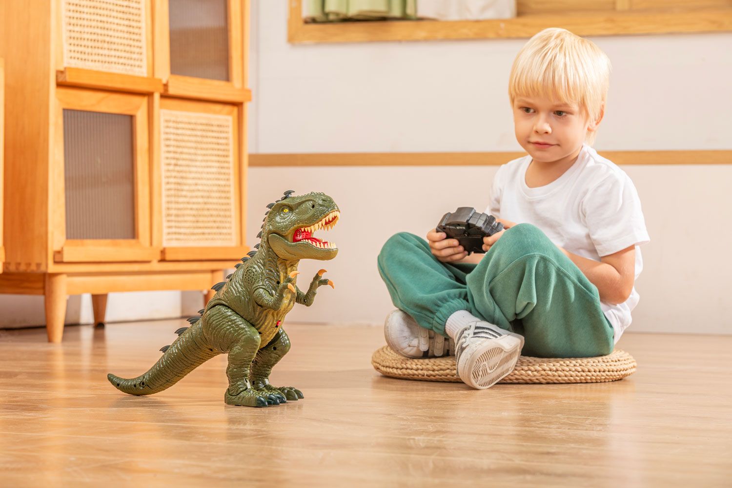 Blonde child seated on a woven mat on the floor with a remote control, looking intently at a toy dinosaur standing upright, showcasing interactive play.