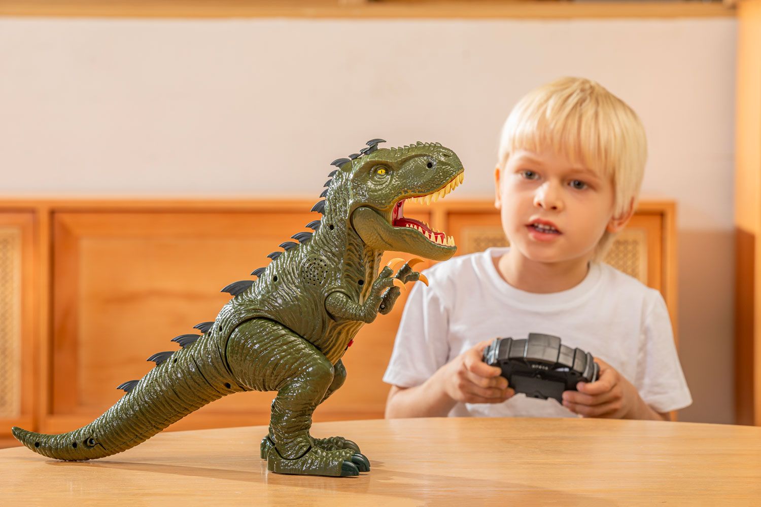 A young blonde child holding a remote control, with a fierce toy dinosaur on a wooden table, capturing a moment of imaginative play.